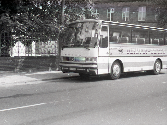 Ein Bus des West-Berliner Unternehmens "Olympia Reisen" parkt am Straßenrand vor einem Mauerzaun, hinter dem ein Backsteingebäude zu erkennen ist.
