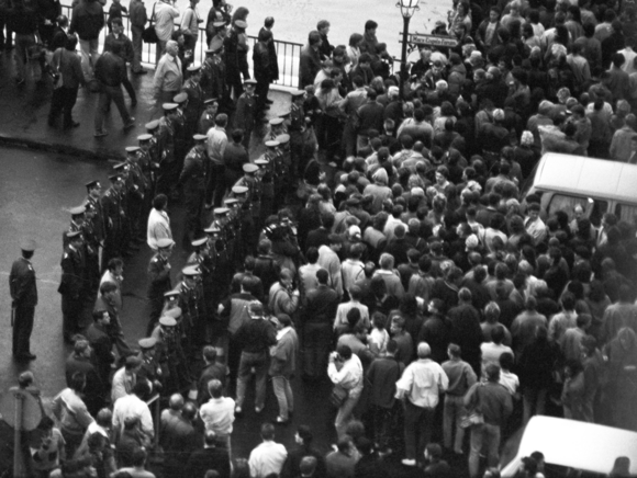 [Das schwarz-weiße Lichtbild zeigt auf der rechten Seite eine Menschenmasse auf der Straße, dazwischen parkende Autos. Aufder linken Seite stehen Polizisten in Doppelreihe zur Abgrenzung. Zwischen ihnen vermutlich Mitarbeiter der Staatssicherheit.]