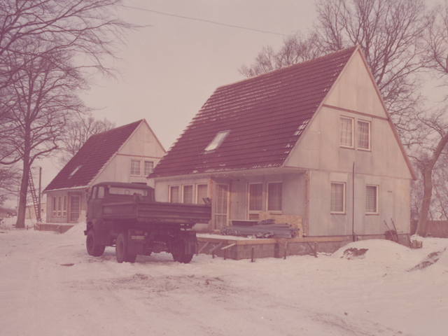 [Das Foto zeigt die diagonale Frontansicht zweier nebeneinander angeordneter, baugleicher zweigeschossiger Häuser mit Spitzdach. Die Fassaden sind noch unverputzt, die Dächer rot geziegelt. Der Boden, umliegende Sandhügel und zum Teil auch die Dächer sind mit Schnee bedeckt. Auf der Terrasse des rechten Hauses liegen aufgestapelte Rohre, Holz- und andere Bauteile. An der Seitenwand lehnt eine Schubkarre. Vor dem Haus parkt ein dunkelgrüner LKW mit Ladefläche.]
