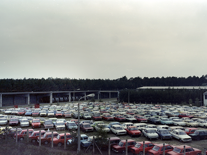 Auf einem unbefestigten, eingezäunten Platz stehen in verschiedenen Modellen und Farben Pkws, die dicht an dicht in Schrägstellung abgeparkte wurden. Auch ein Wohnwagen steht am Rand. Im Hintergrund sind auf der rechten Seite lang gezogene Flachbauten zu erkennen, auf der linken Hälfte grenzt ein Wald an. An den Zäunen stehen Laternen. Es handelt sich um ein farbiges Lichtbild, das erhöht mit einem Weitwinkelobjektiv aufgenommen wurde.