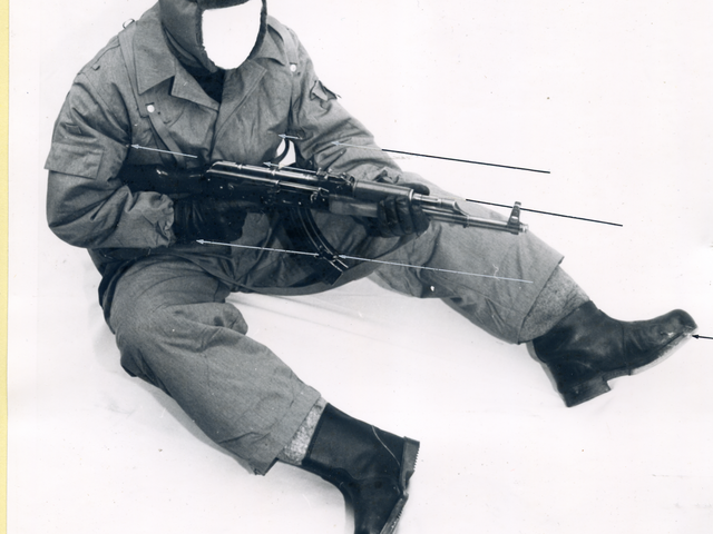 [Ein schwarzweißes Lichtbild, das eine sitzende Personen in wattierter Winteruniform der Grenztruppen inklusive Fellmütze zeigt. Es soll eine Nachstellung der möglichen Haltung von Seidel zur Schussabgabe darstellen. Die Person wurde bereits durch das MfS anonymisiert, statt einem Gesicht ist ein Feld des hellem Hintergrund eingefügt. Dargestellt wird eine sitzende Position, in der beide Beine fast ausgestreckt sind. Dabei wird ein Maschinengewehr gerade in Anschlag genommen, aber der Lauf ist noch eher gen Boden geneigt. Schwarze und graue Pfeile sind exakt eingezeichnet worden.]