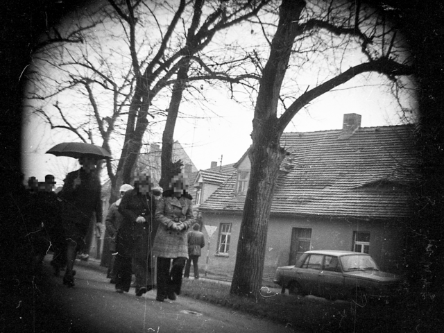 Mehrere zumeist dunkel gekleidete Personen gehen eine vermutlich ländlich gelegene Straße entlang. Im Hintergrund sind Bäume, Wohnhäuser und ein parkender Wartburg zu sehen.