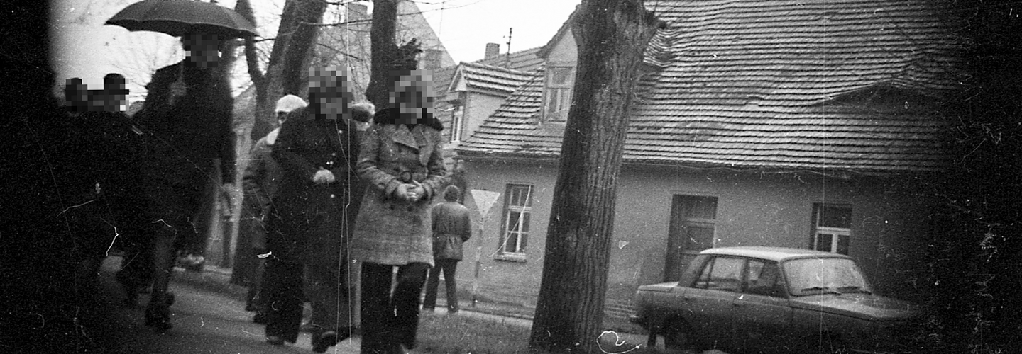 Mehrere zumeist dunkel gekleidete Personen gehen eine vermutlich ländlich gelegene Straße entlang. Im Hintergrund sind Bäume, Wohnhäuser und ein parkender Wartburg zu sehen.
