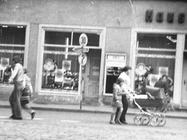 [Bei der Abbildung handelt es sich um ein schwarz-weißes Negativ. Von einem gepflasterten Platz aus wurde die Schaufensterzeile eines älteren Hauses auf der anderen Straßenseite fotografiert. Die Außenreklame ist nur anteilig "Hauss" abgebildet. Es sind einige Passanten unterwegs, darunter auch Kinder. Die Aufnahme wurde vermutlich verdeckt aufgenommen, der Bildausschnitt wird rund und die Ecke abgedunkelt.]