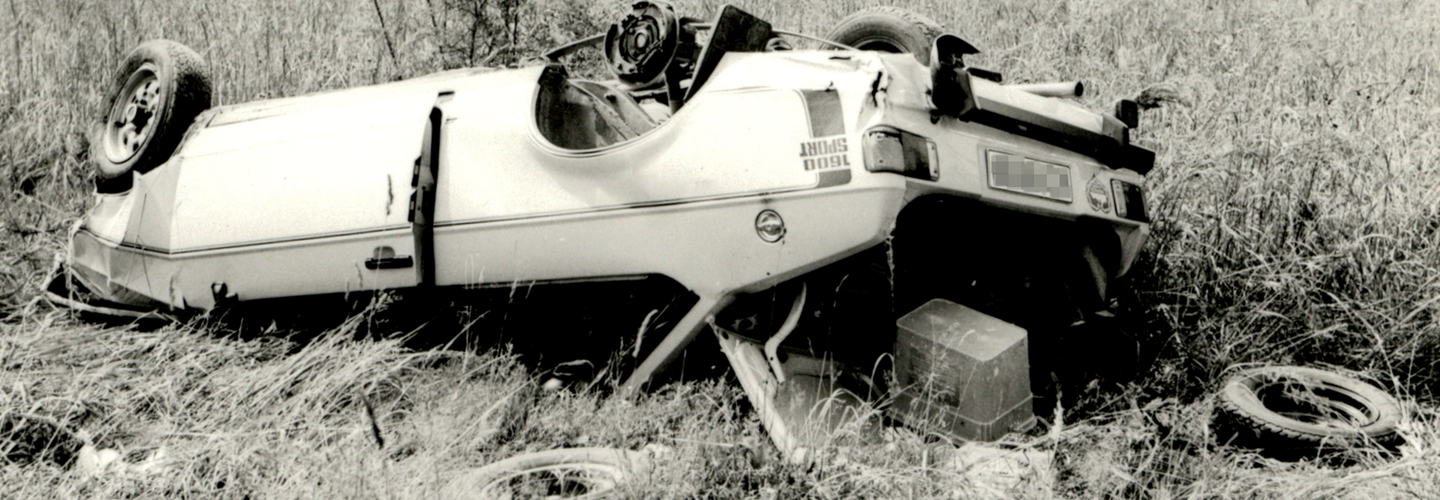Das stark beschädigte Unfallfahrzeug liegt abseits der Straße auf dem Dach. Daneben sind Reifen und der Inhalt des geöffneten Kofferraums auf der Wiese verstreut.
