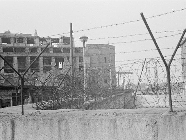 Der Fokus des schwarz-weißen Lichtbilds liegt auf dem Stacheldraht auf einem Mauerabschnitt. Im Hintergrund sind drei Gebäudeschuppen und ein großes, vom Krieg geschädigtes Haus zu sehen.