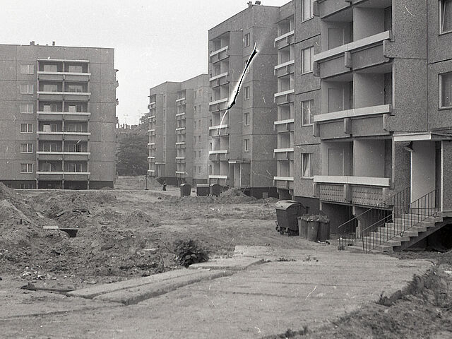 [In mitten mehrerer sechsstöckiger Wohnblocks (vermutlich Wohnbauserientyp 70) ist aufgewühlter Boden zu sehen. Diese baustellenartig-wirkenden Erdhaufen nehmen einen Großteil des Bildes ein. Im vorderen Bildgrund ist ein Weg aus großen länglichen Betonplatten. Es handelt sich um ein schwarzweißes Negativ, das in der Mitte des oberen Drittels einen Einschnitt vorweist.]
