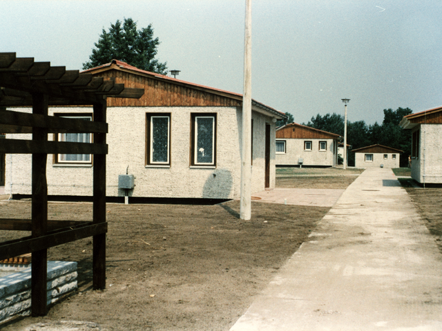 Das Bild zeigt einen Weg, links und rechts davon zweigen weitere Wege ab, die jeweils zu einem Bungalow führen. Links im Vordergrund ist der Teil eines offenen Holzgebäudes zu sehen.