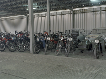Ein mittels Wellblech überdachter Platz aus Betonplatten wird als Parkplatz genutzt. Vor einer Wand aus Wellblech stehen zahlreiche Motorräder in zwei Reihen in senkrechter Aufstellung. In der hinteren Reihe stehen auch drei Trabis. auf der rechten Seite stehen in der vorderen Reihe auch ein Anhänger, beladen mit Papiertüten, sowie links davon ein Motorrad mit Beiwagen. Das farbige Lichtbild wurde mit einem Weitwinkelobjektiv aufgenommen.