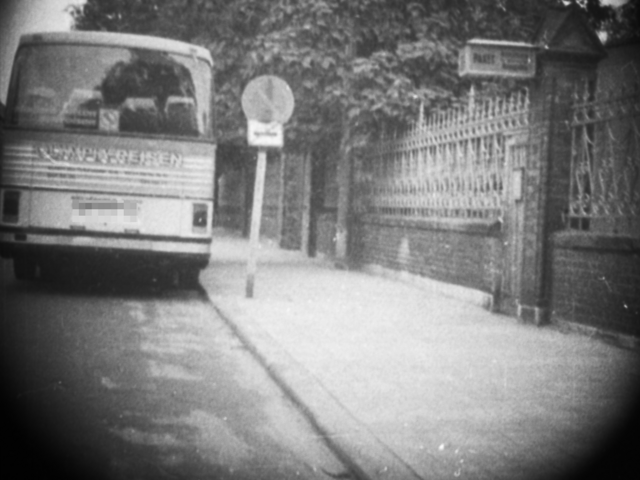 Ein Bus des Westberliner Reiseunternehmens "Olympia Reisen" parkt am Straßenrand neben einem Mauerzaun, direkt hinter einem Halteverbotsschild. Ein Kasten für Leuchtreklame weist darauf hin, dass sich hinter dem Zaun eine Gaststätte befinden könnte.