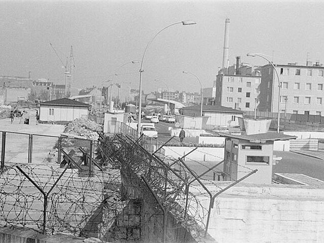 Das schwarz-weiße Lichtbild wurde oberhalb des Stacheldrahtes der Mauer aufgenommen. Zu sehen ist die Grenzübergangsstelle Heinrich-Heine-Straße mit Blick gen Berlin (Ost). Auf der linken Seite liegt ein braches Grundstück, auf dem Schuttberge zu aufgehäuft sind.