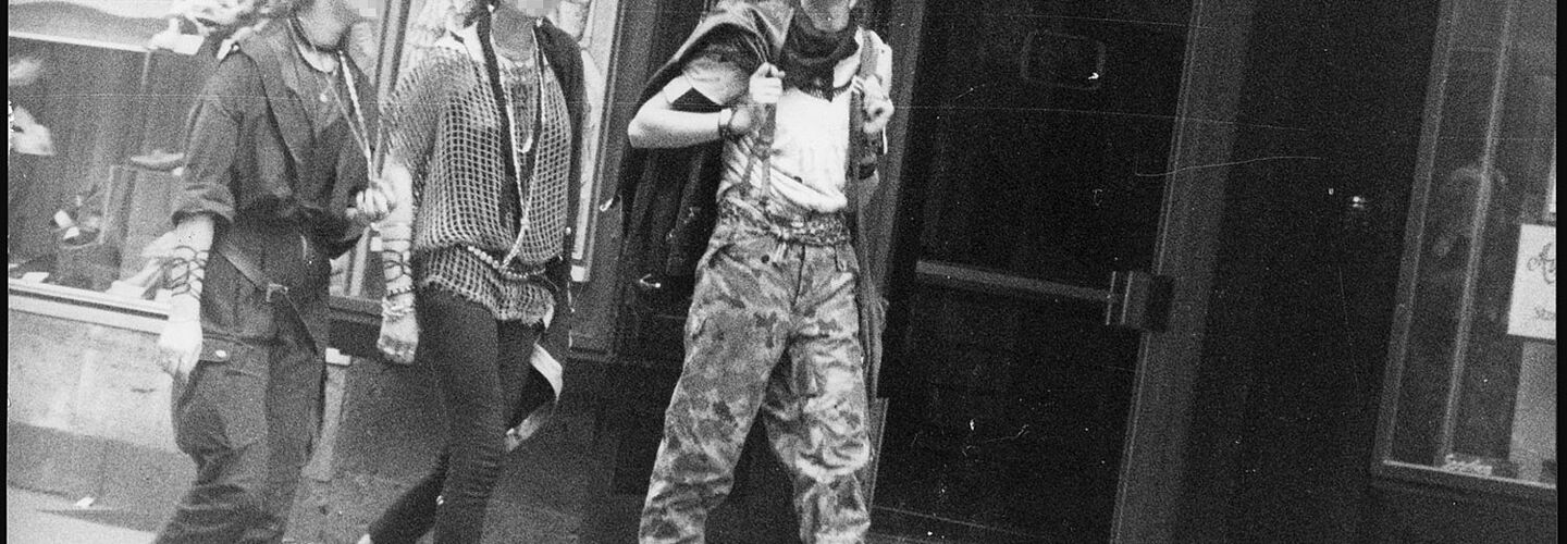 Schwarz-Weiß-Foto mehrerer Personen im Punk-Look auf einer Straße, die an der anscheinend versteckten Kamera vorbei laufen.