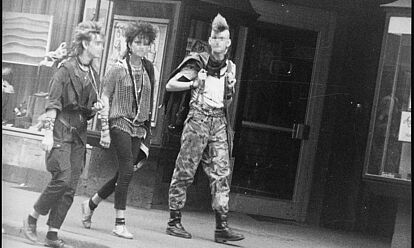 Schwarz-Weiß-Foto mehrerer Personen im Punk-Look auf einer Straße, die an der anscheinend versteckten Kamera vorbei laufen.