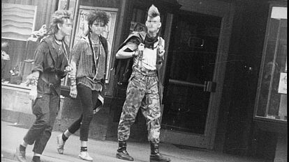 Schwarz-Weiß-Foto mehrerer Personen im Punk-Look auf einer Straße, die an der anscheinend versteckten Kamera vorbei laufen.